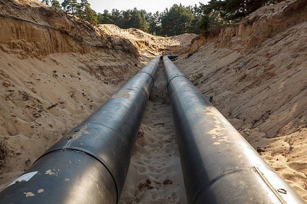 Two long, black industrial pipes lying parallel at the bottom of a sandy, excavated trench in a forested area.