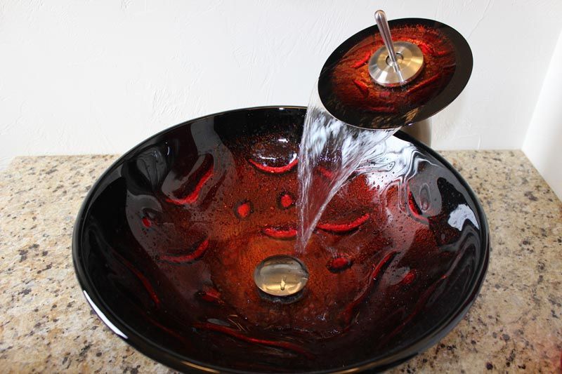 Red glass tabletop fountain-style sink with water pouring into a matching bowl on a stone counter
