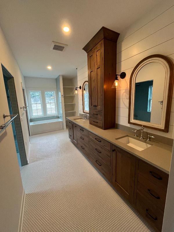 Long bathroom with double vanity, wood cabinetry, wall mirror, and bright windows at the far end.