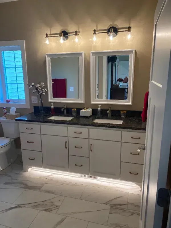 Bathroom vanity with double mirrors, wall lights, gray cabinets, and a marble floor under a window.