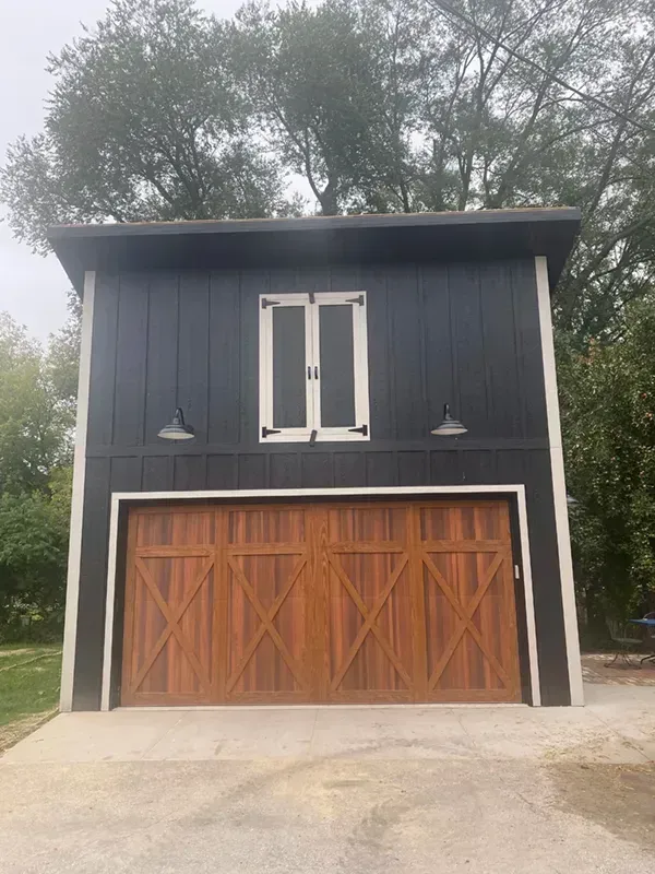 Two-story black garage with wooden double doors and small balcony windows beneath trees