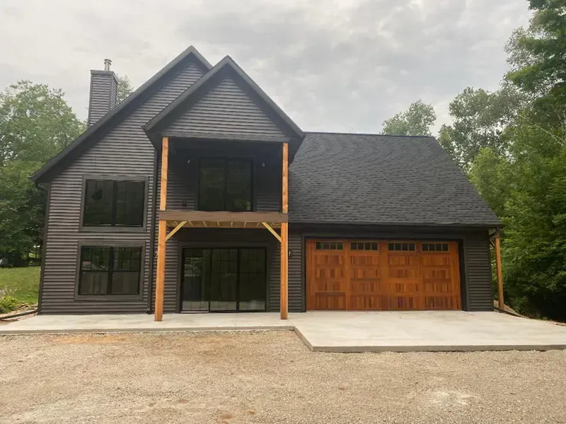 Modern two-story house with dark siding, large windows, and a wooden garage door, viewed from the driveway