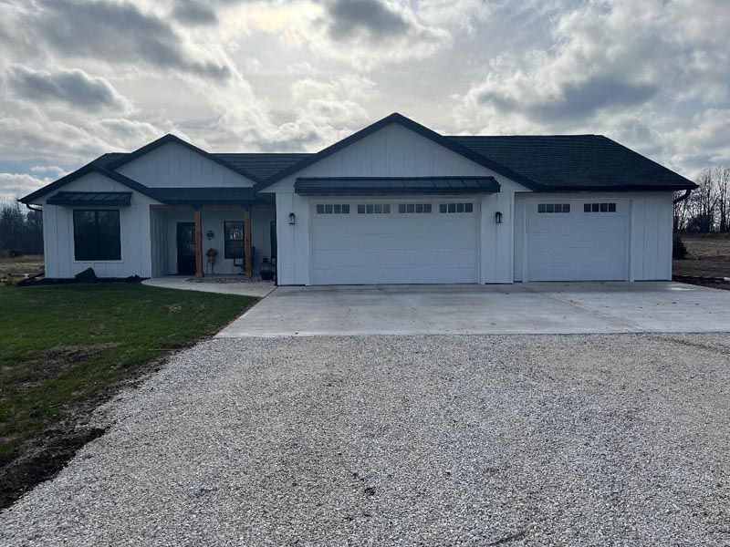 Single-story suburban house with gray roof, white garage doors, and gravel driveway