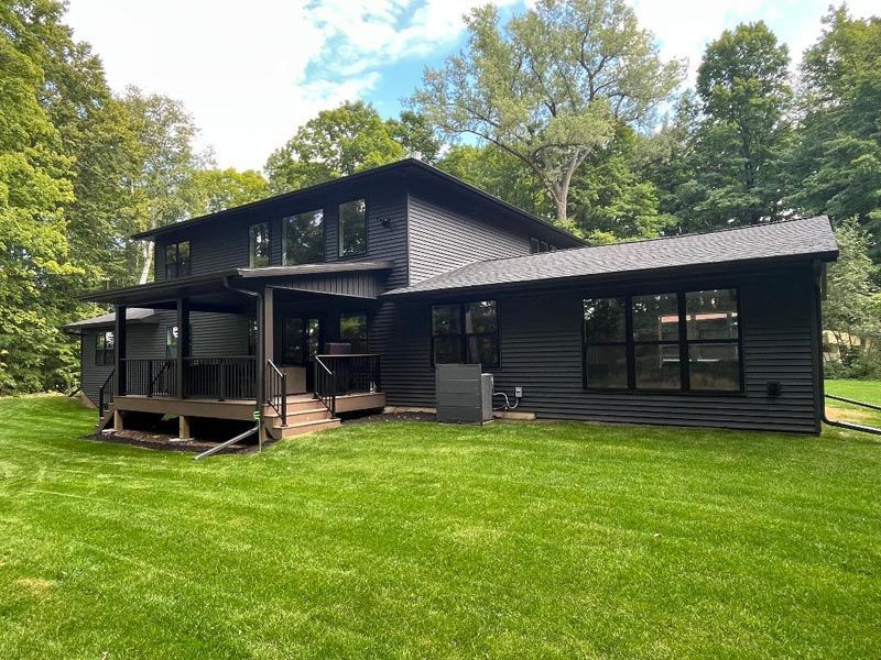 Black two-story house with a covered porch and large lawn, surrounded by trees under a blue sky
