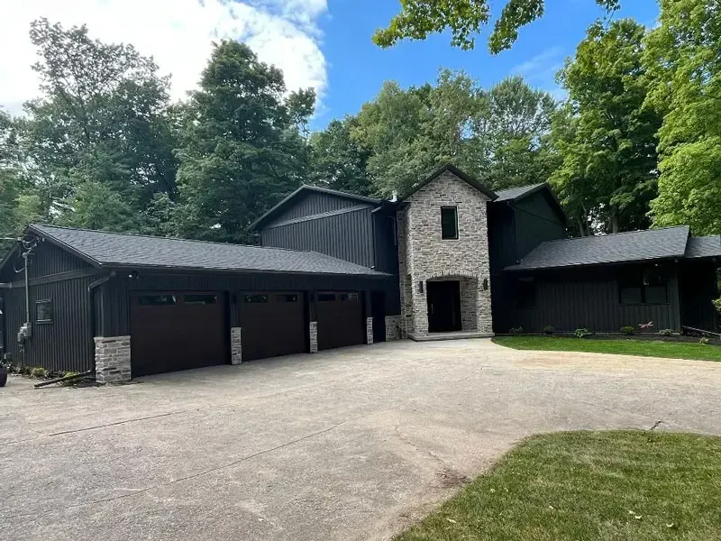 Large modern house with stone entry, dark siding, and a wide driveway surrounded by trees