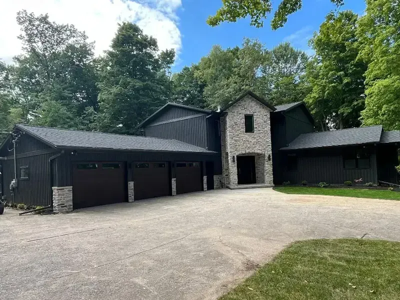 Large dark house with stone entry, wide driveway, and trees under a blue sky