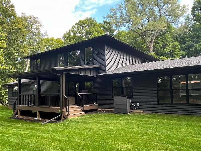 Black modern house with dark siding and a large porch, surrounded by a green lawn and trees.
