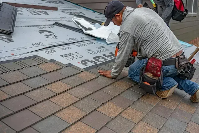 Worker installing brown roof shingles beside gray underlayment on a rooftop