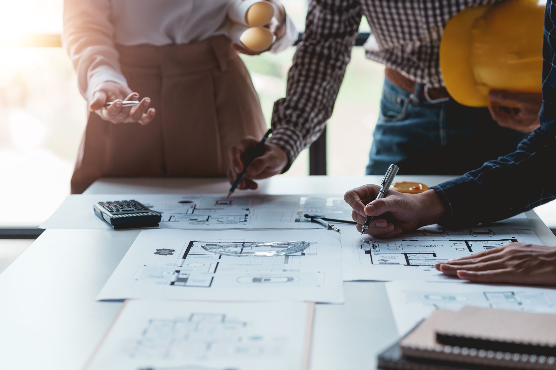 People reviewing architectural blueprints at a table, with laptop and coffee in a bright office.