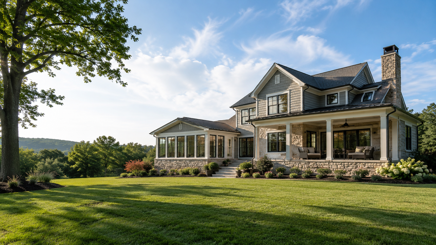 Large stone farmhouse with wraparound porch on a sunny lawn, framed by trees and hills.