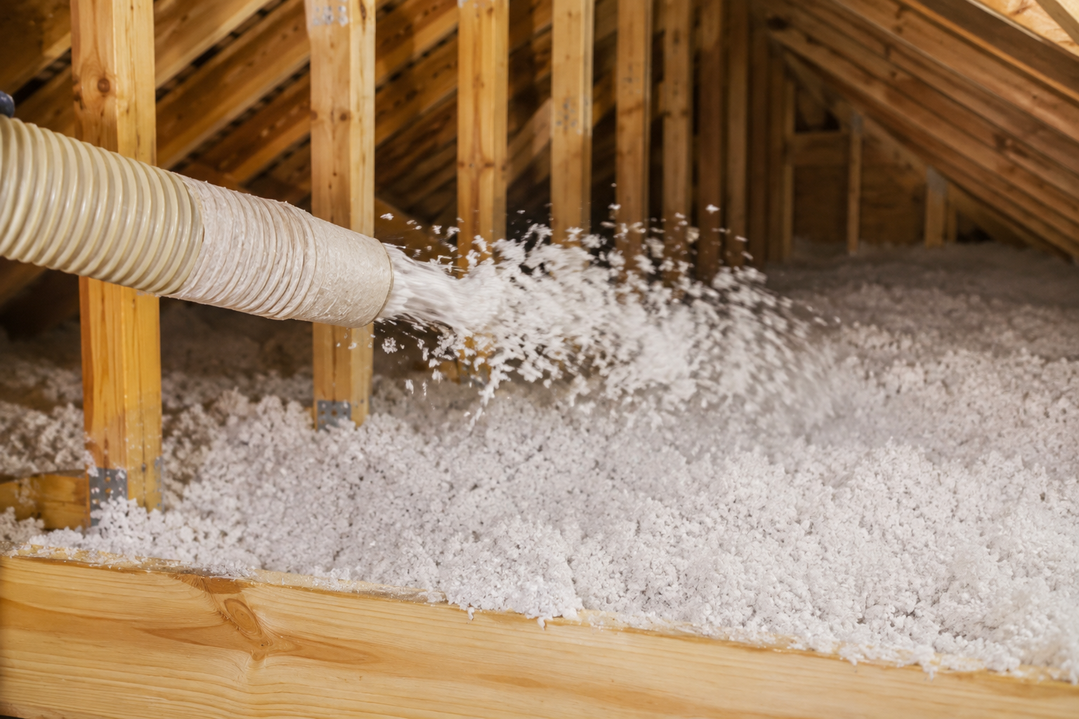 Attic insulation blown through a hose into the rafters, with white cellulose piling on the floor