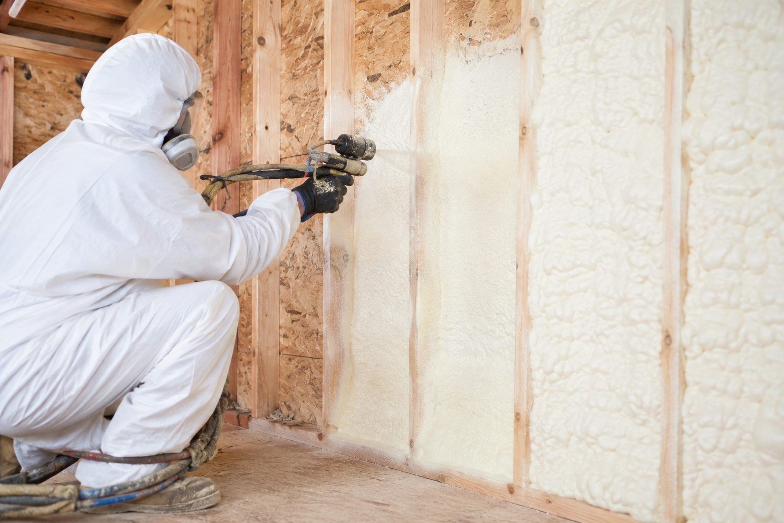 Worker in white protective suit spraying foam insulation on a wall inside a wooden-framed room