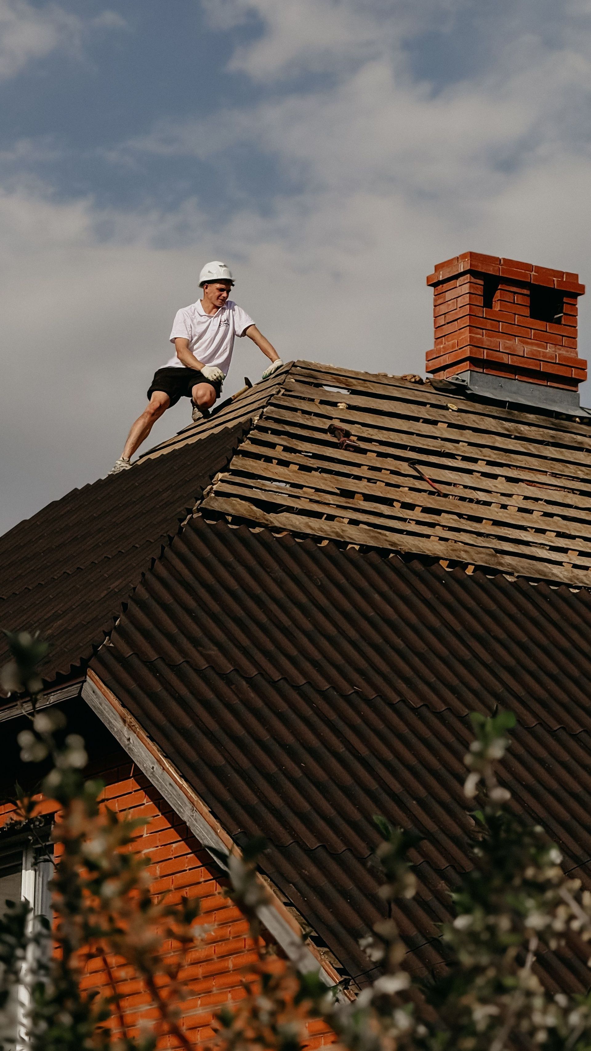 Roofer in white shirt and helmet working on a sloped roof beside a brick chimney