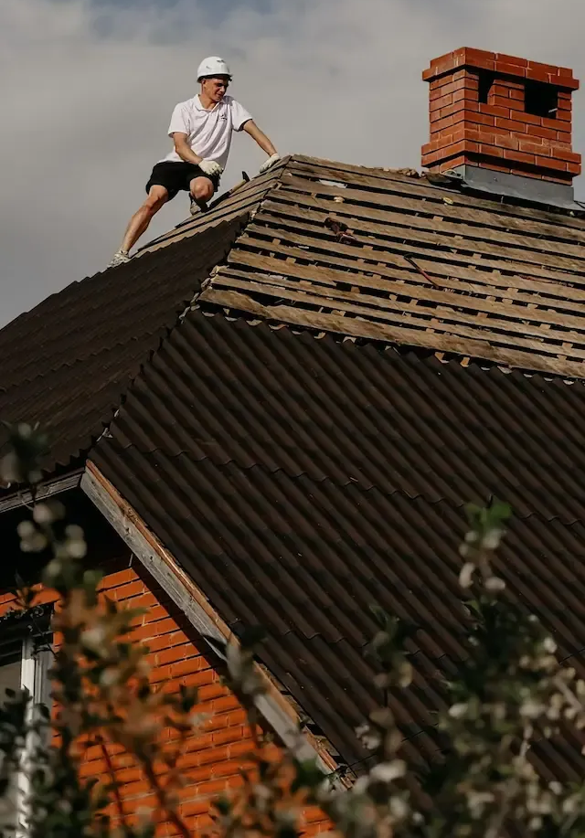 Worker in white hard hat crouches on a roof beside a red brick chimney.