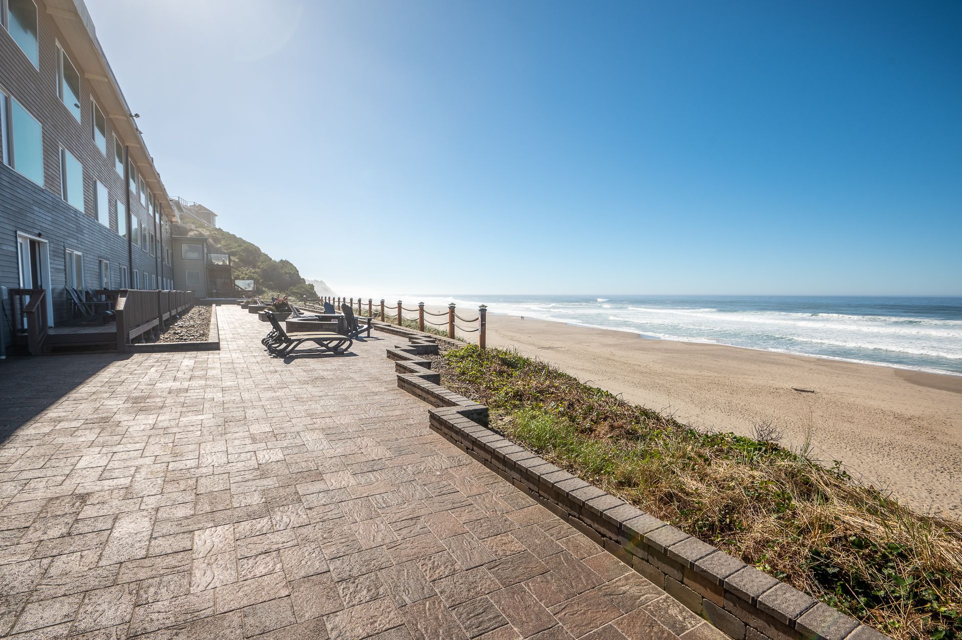 A stone patio with outdoor seating overlooks a sandy beach and the ocean under a clear, bright blue sky.