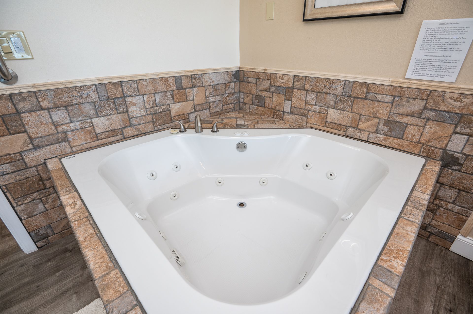 A corner jetted bathtub with a white basin, surrounded by rustic stone-patterned tile walls and countertop.