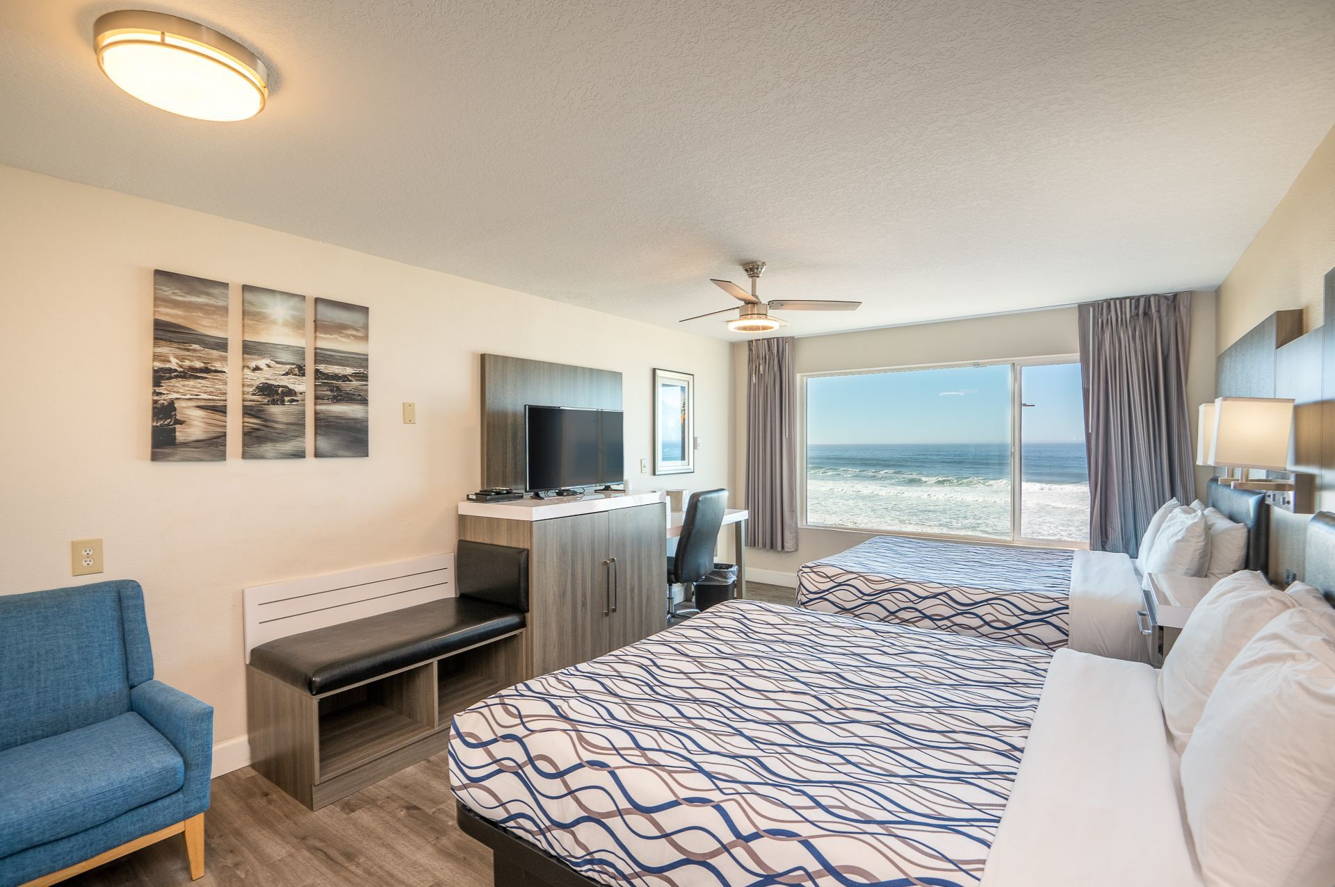 A hotel room with two queen beds, a blue armchair, a television on a dresser, and a large window overlooking the ocean.