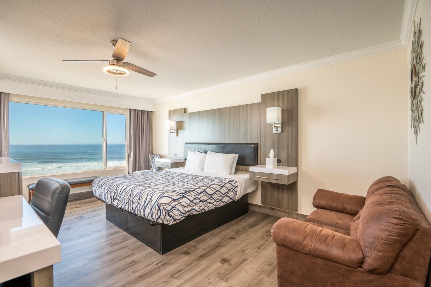 Hotel room with ocean view, bed, sofa, desk, and ceiling fan.