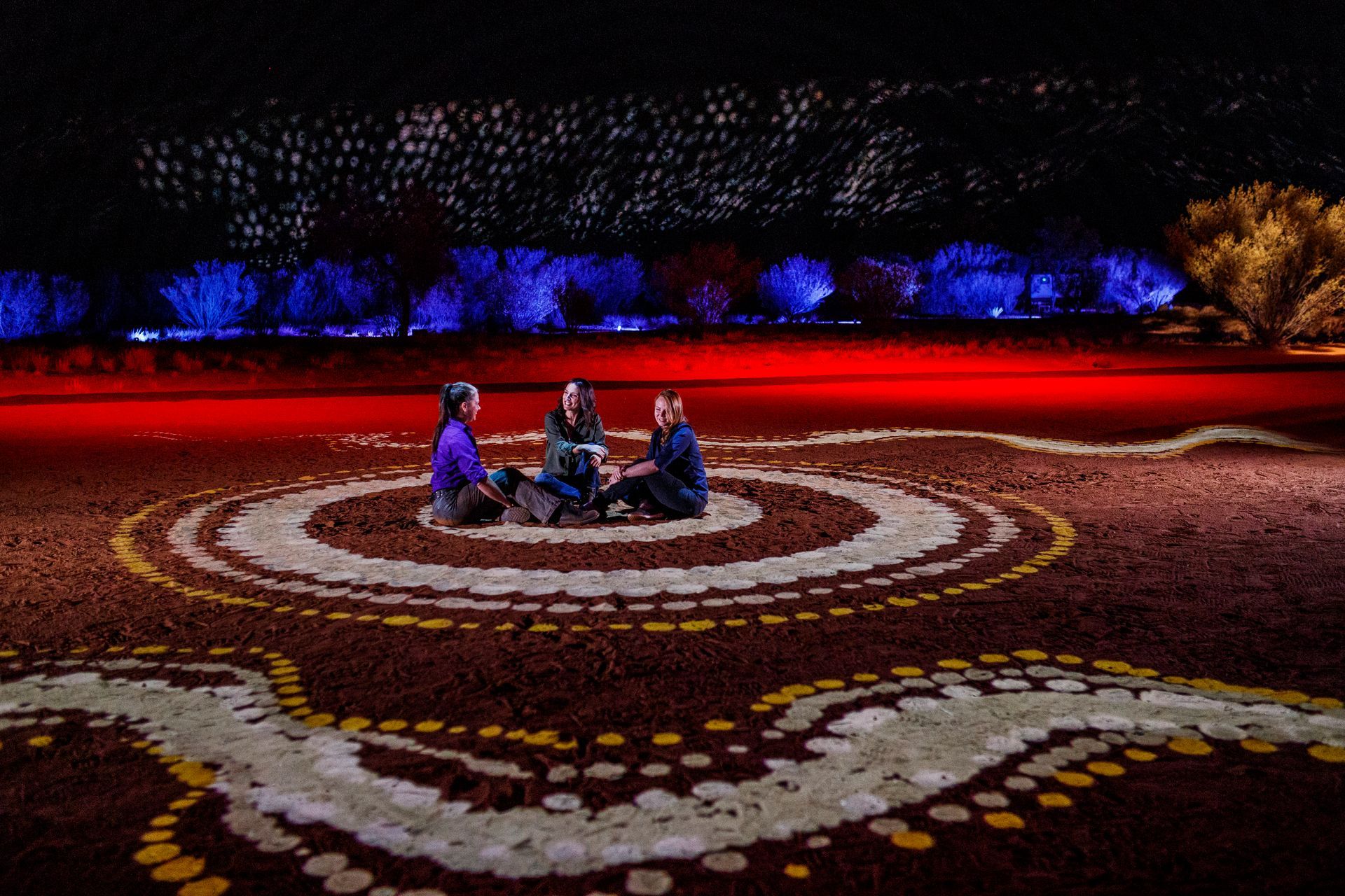 Three people sit in a circle on patterned white stones illuminated on red earth, with blue lighting in the background.