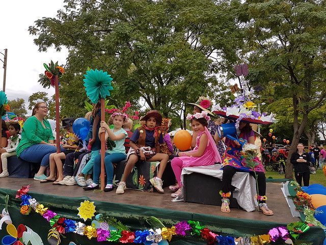 A group of children in colorful costumes riding on a decorated parade float outdoors with trees in the background.