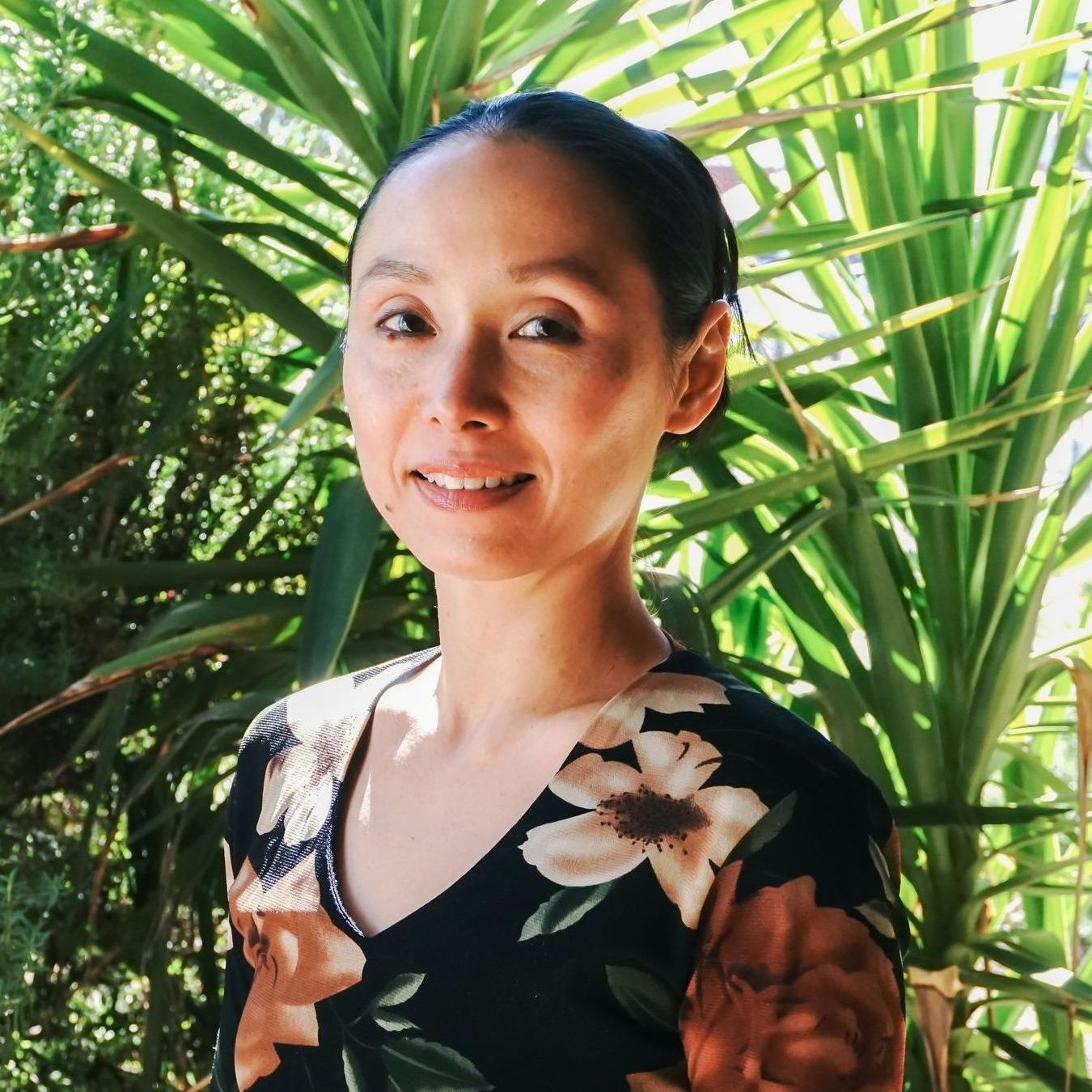 A person with hair pulled back smiles gently while standing outdoors in front of lush green plants.