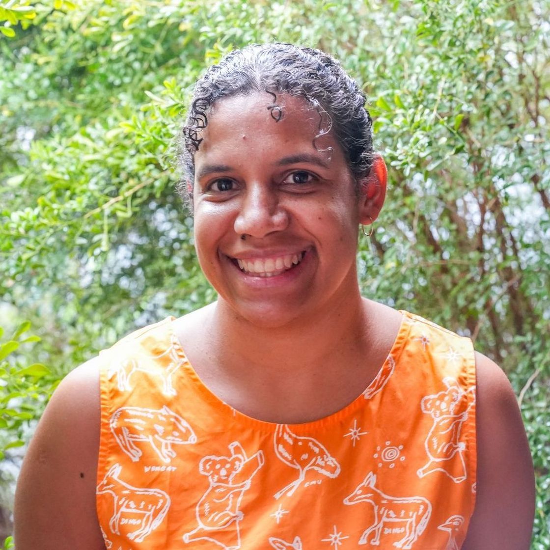 A smiling person wearing an orange shirt with white animal sketches, standing in front of lush green foliage.