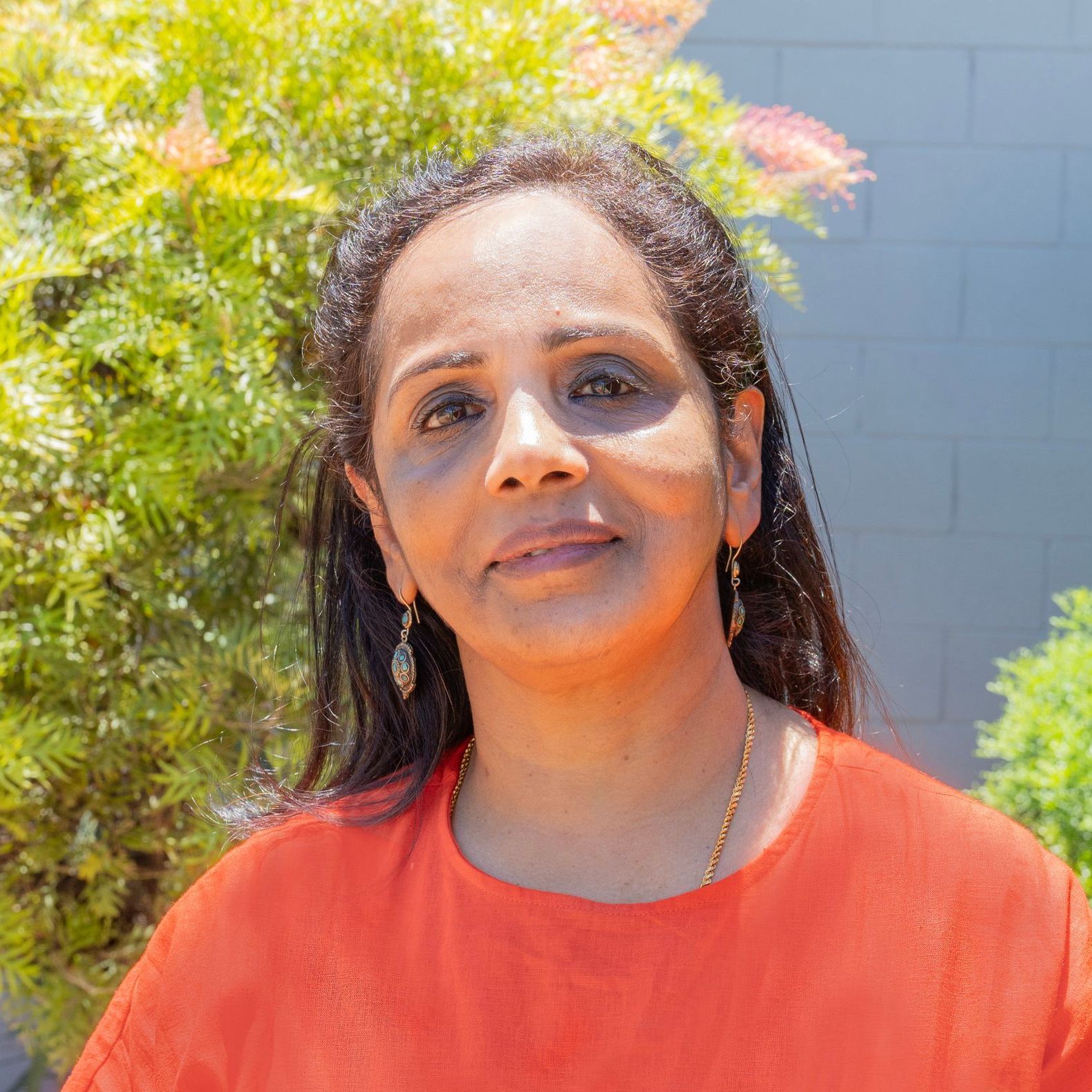 A woman with long, dark hair wears a bright orange top and gold earrings, posing in front of lush green foliage.