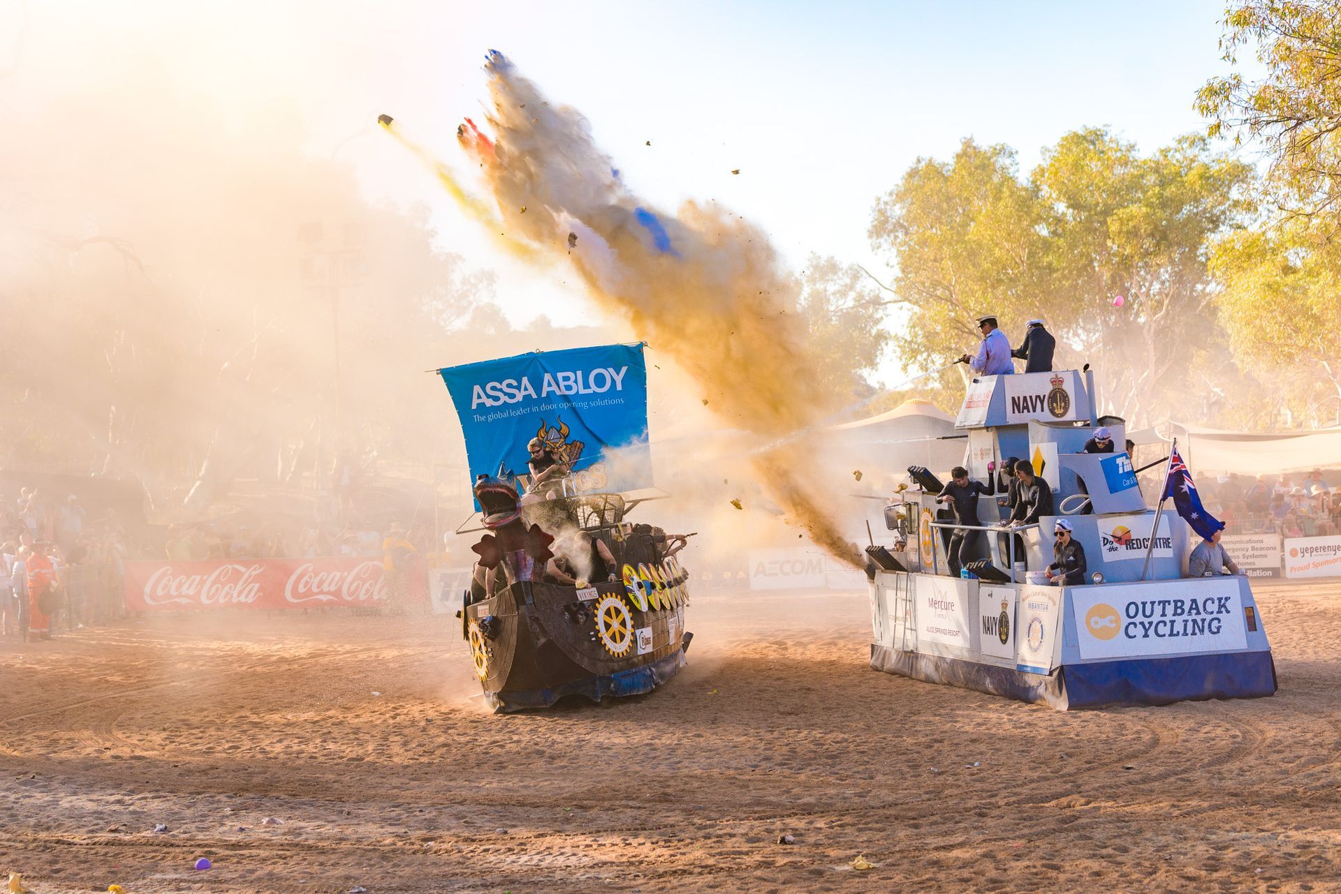 Two festive, decorated vehicles in a sandy arena engage in a playful mud-spraying battle under bright sunlight.