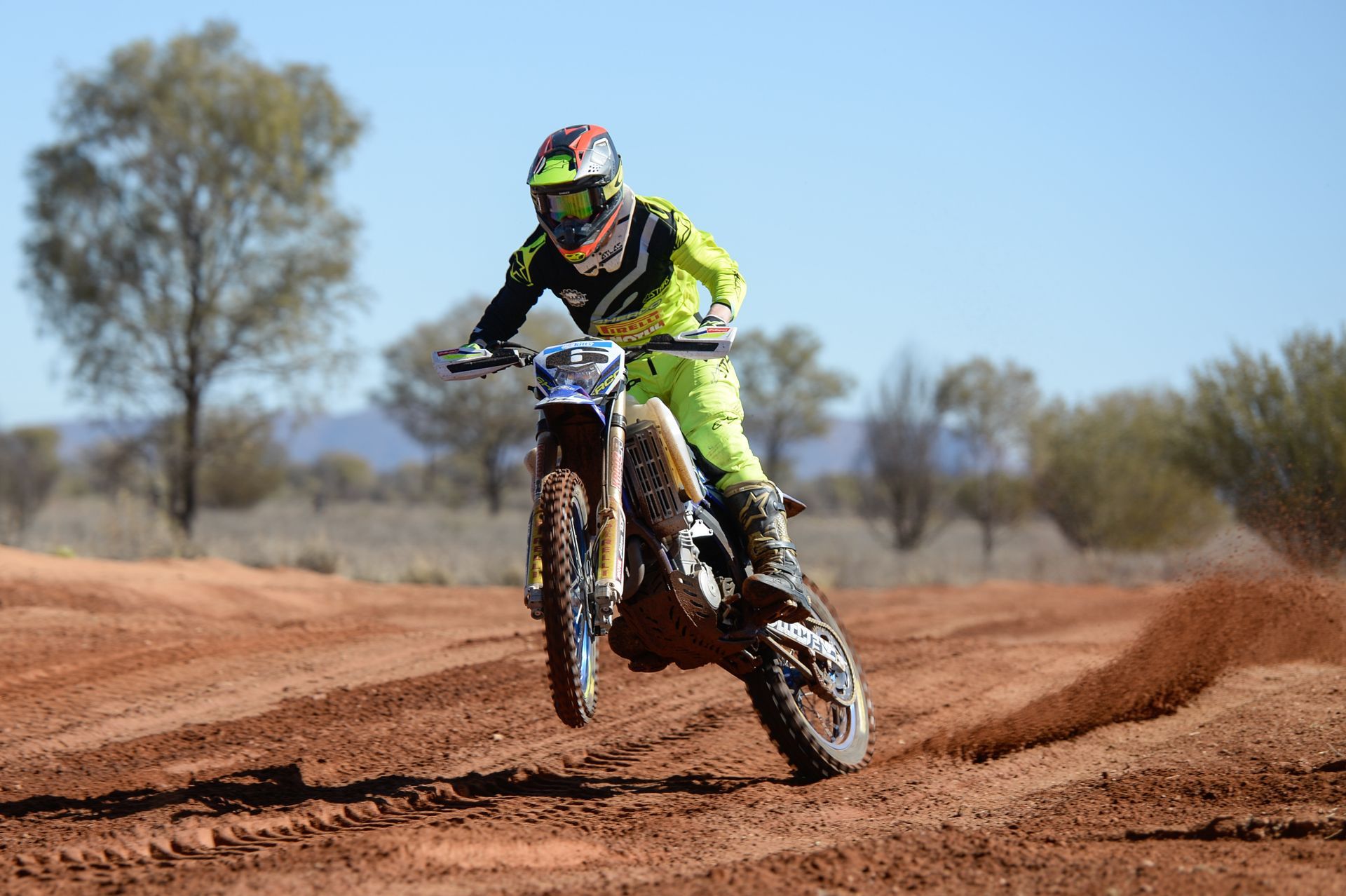 A dirt bike rider in neon yellow gear performs a jump on a red dirt track under a clear blue sky.