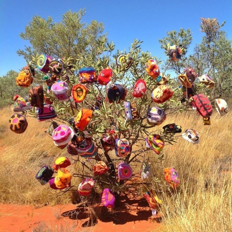 A desert shrub decorated with many colorful, patterned crocheted and knitted hats, set against a bright blue sky.