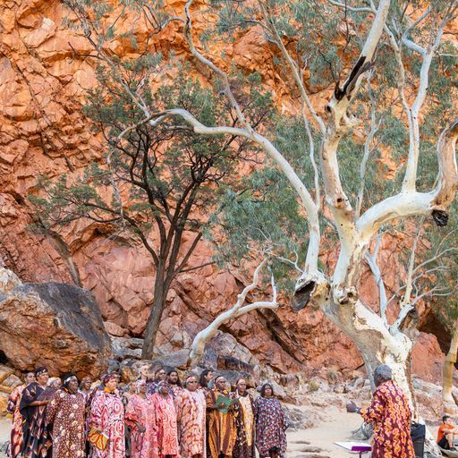 A group in patterned tunics performs outdoors before a red rocky cliff and large ghost gum trees.