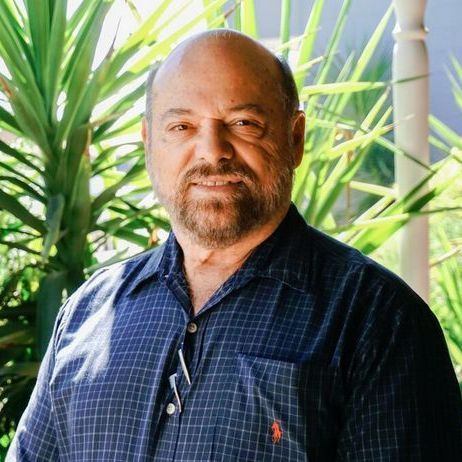 A person with a beard, wearing a dark blue plaid button-down shirt, poses in front of green palm leaves.