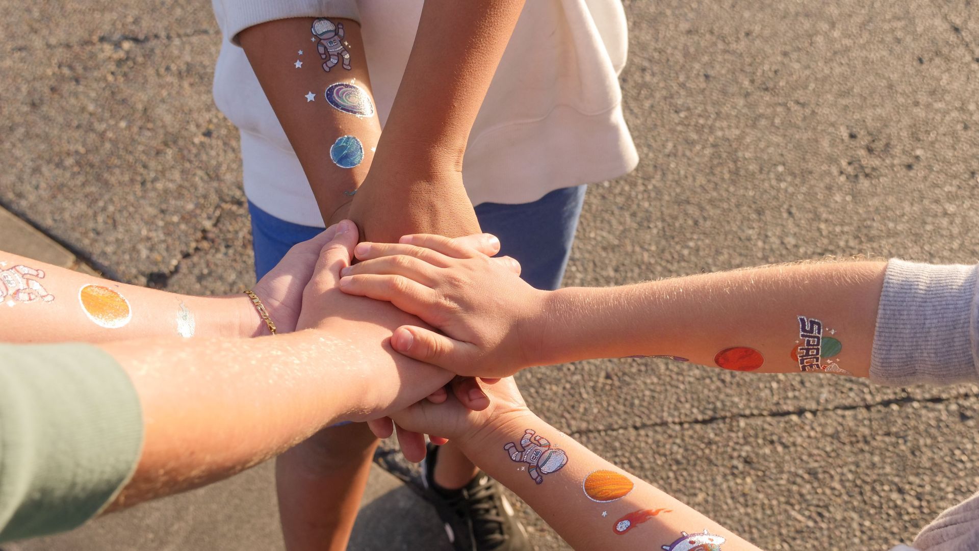 Several people stack hands together outdoors on pavement, arms showing colorful stickers.