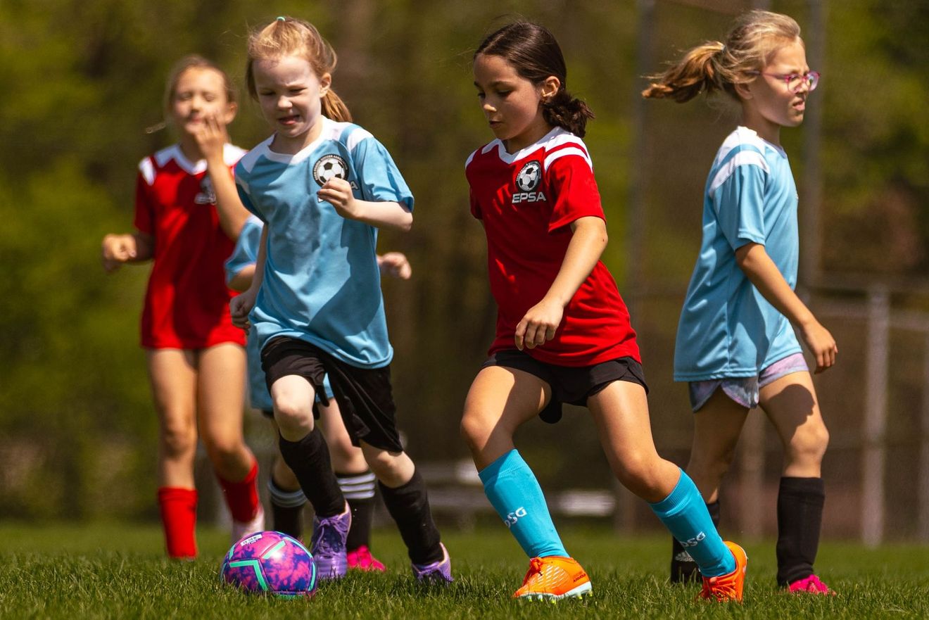 Girls playing soccer on a grass field: two in light blue, two in red, with a purple and black ball.