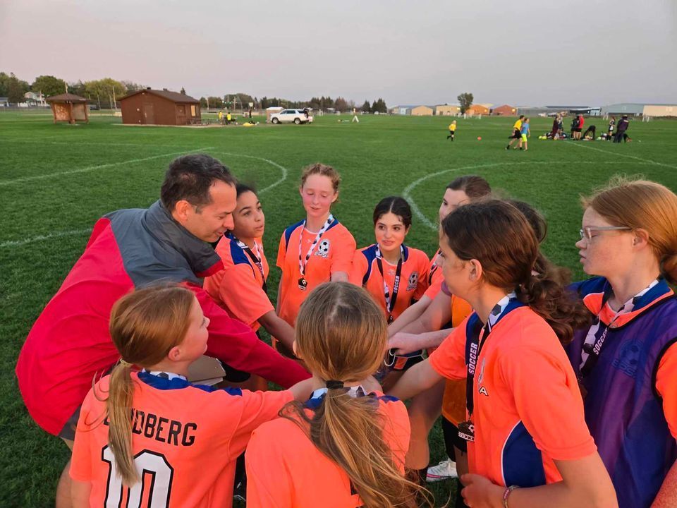 Soccer team huddle with coach on field in orange jerseys, hands in the center.