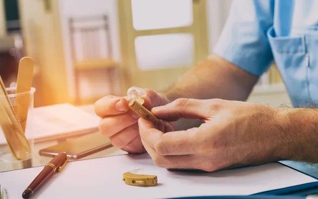 A man is sitting at a desk holding a hearing aid in his hands.