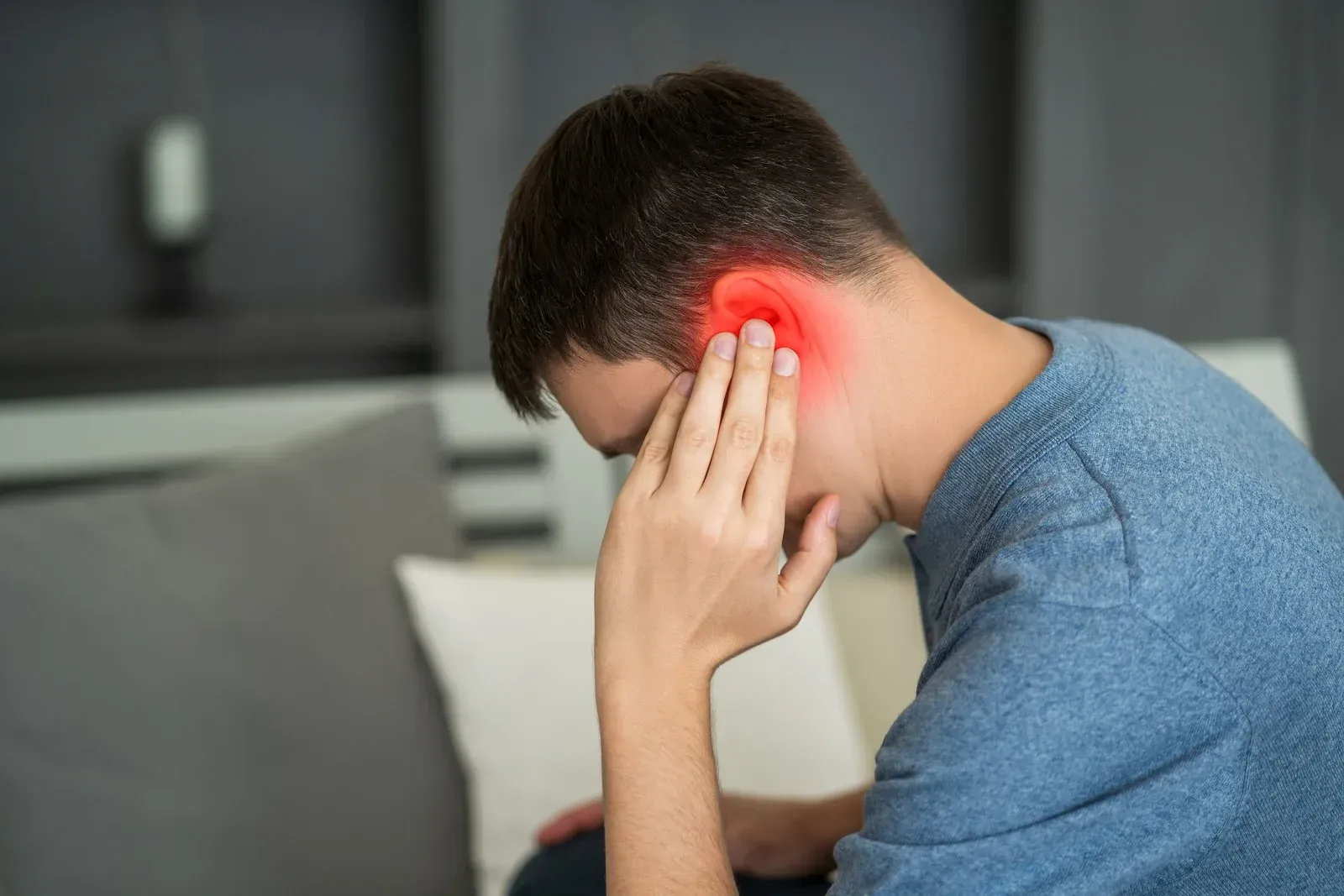 A man is sitting on a couch holding his ear because he has a headache.