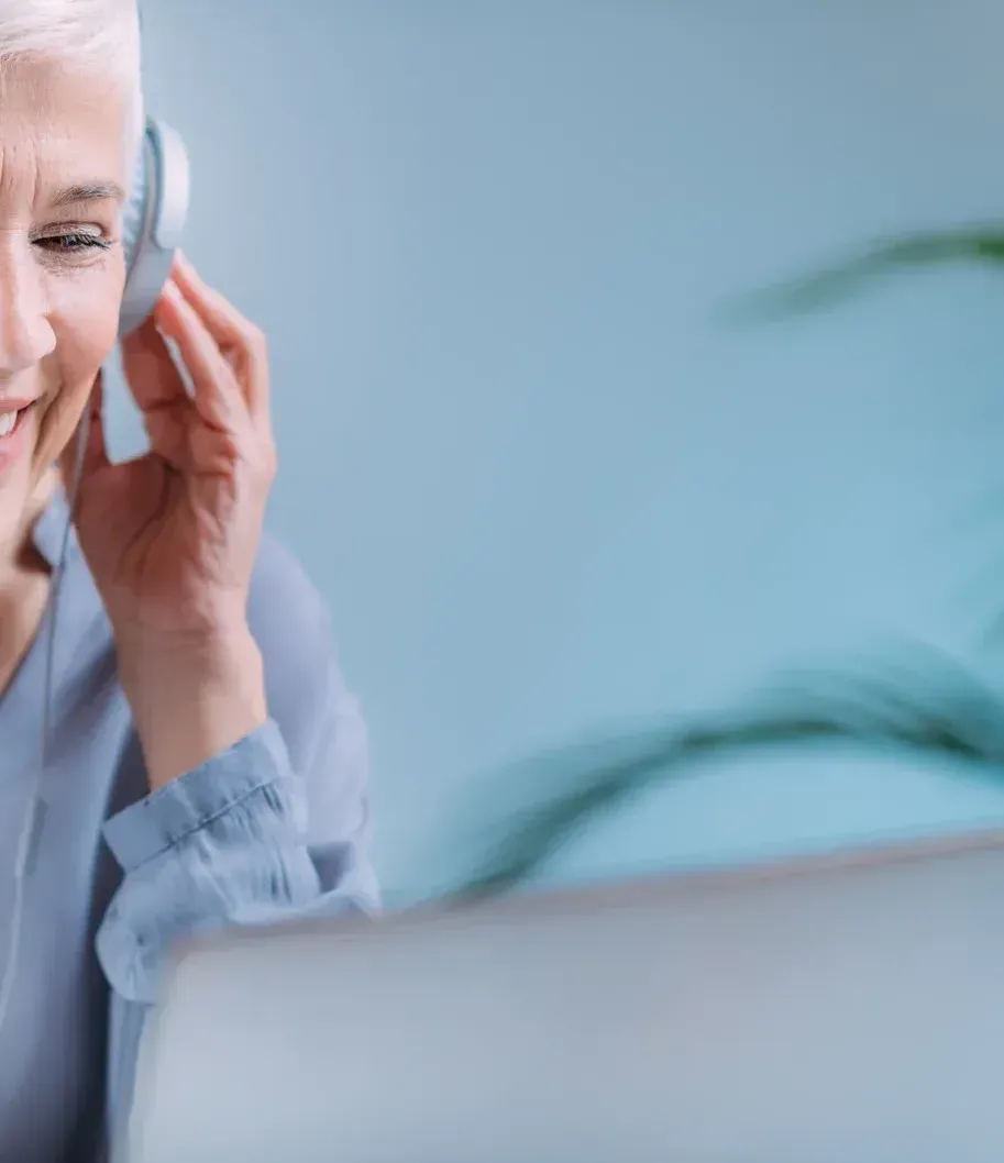 An older woman is wearing headphones while using a laptop computer.