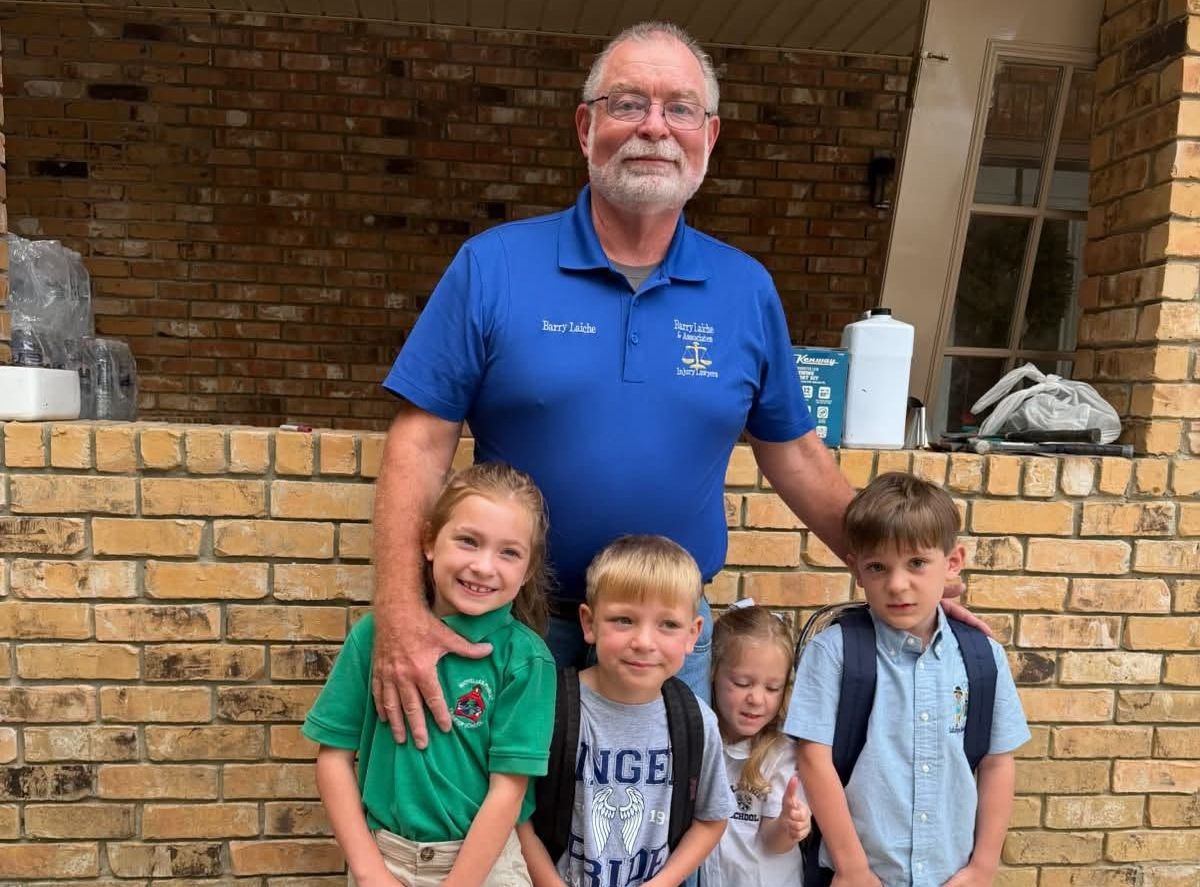 Man in blue shirt standing behind three smiling children in front of a brick wall and porch.
