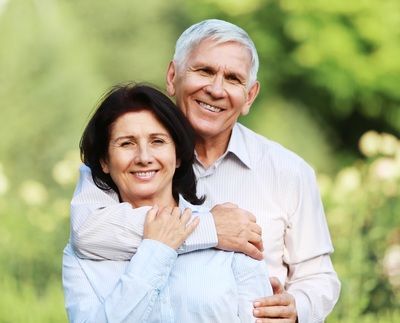 A smiling couple standing outdoors in a green, softly blurred garden, with one person embracing the other from behind.