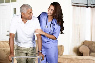 A healthcare professional assists a person walking with a mobility aid in a home setting.