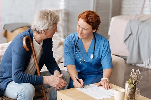 A healthcare worker in blue scrubs sits with a person, completing paperwork on a table in a home setting.