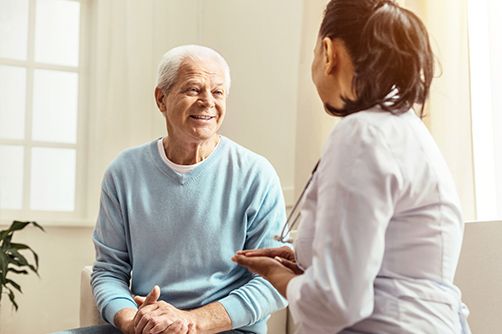 A healthcare provider in a white coat speaking with a smiling patient in a blue sweater in a brightly lit room.