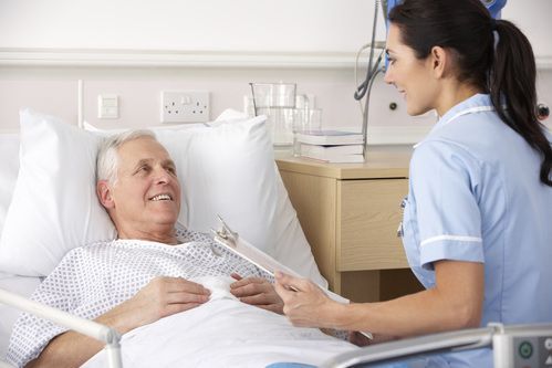 A healthcare worker in a blue uniform reviews a clipboard with a smiling patient resting in a hospital bed.