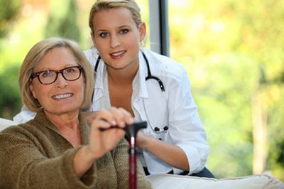 A healthcare professional stands behind a smiling person holding a cane in a brightly lit room.