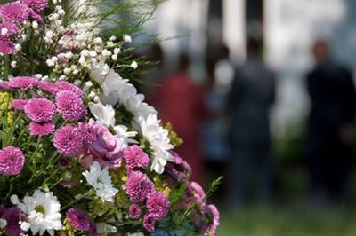 A floral arrangement of purple chrysanthemums and white flowers in the foreground, with blurred figures standing behind.