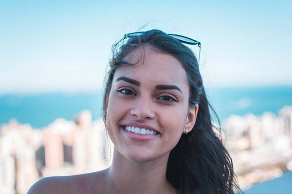 A smiling person with dark hair and sunglasses worn on their head, set against a blurred background of a coastal city.