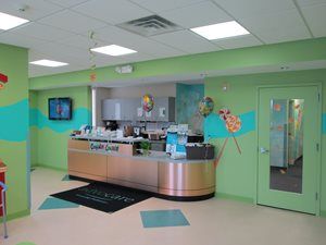 Bright green pediatric reception desk with balloons, TV, and double doors in a clinic lobby
