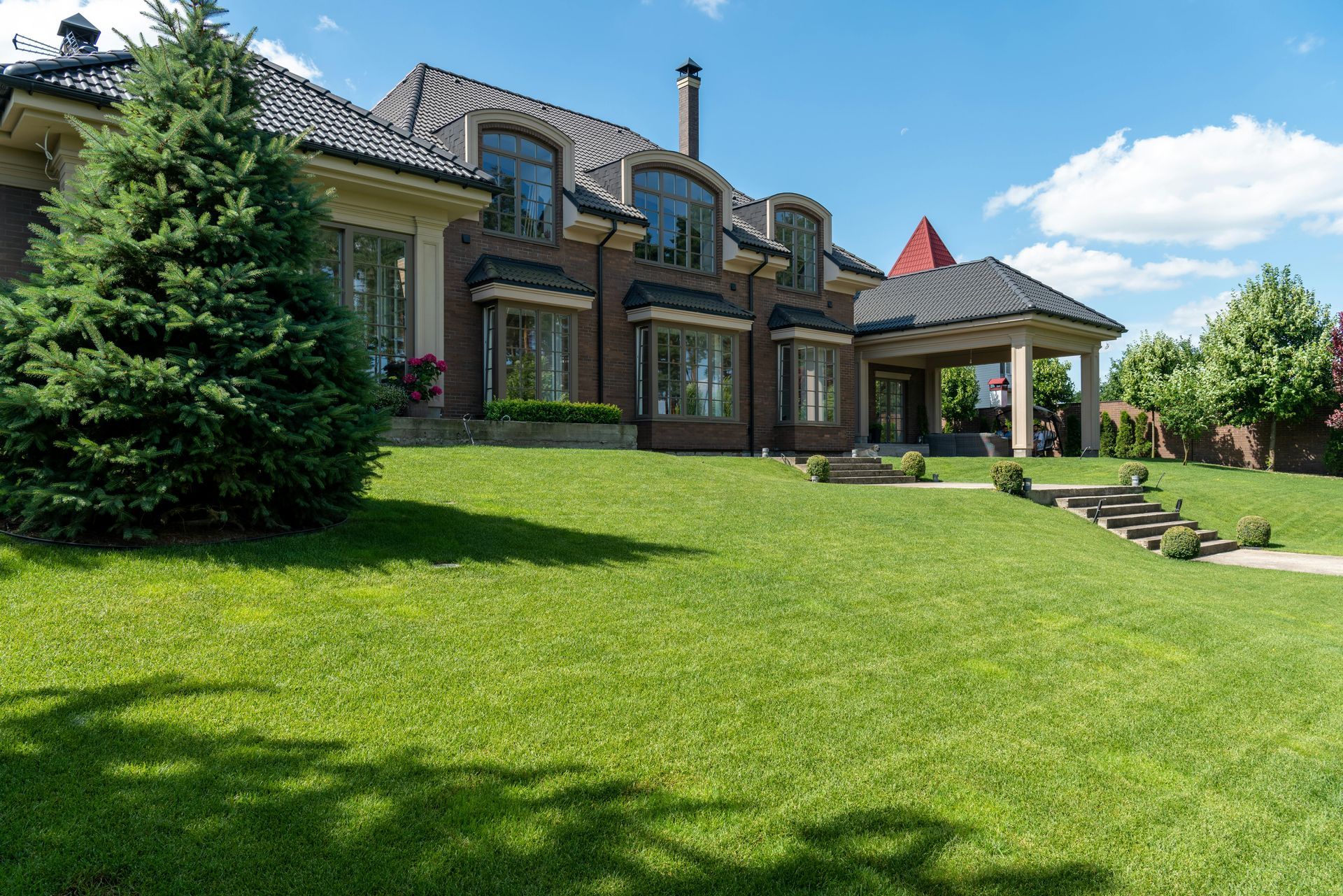 Large brick house with tiled roof, covered porch, and a wide green lawn under a clear blue sky.