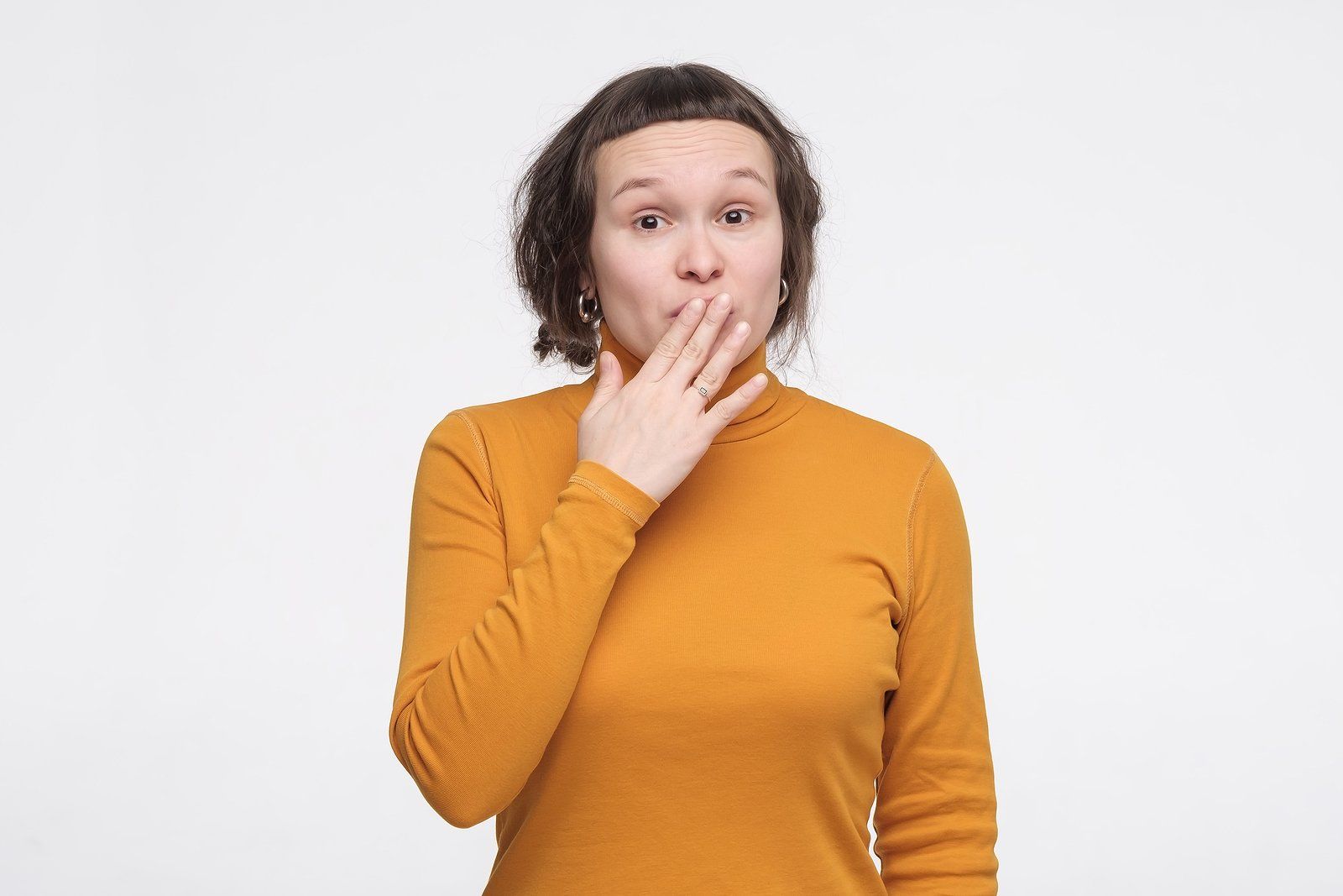 Person in an orange shirt covering their mouth with a surprised expression against a white background